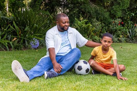 African american father and son bonding in backyard with soccer ball, relaxing on lawn