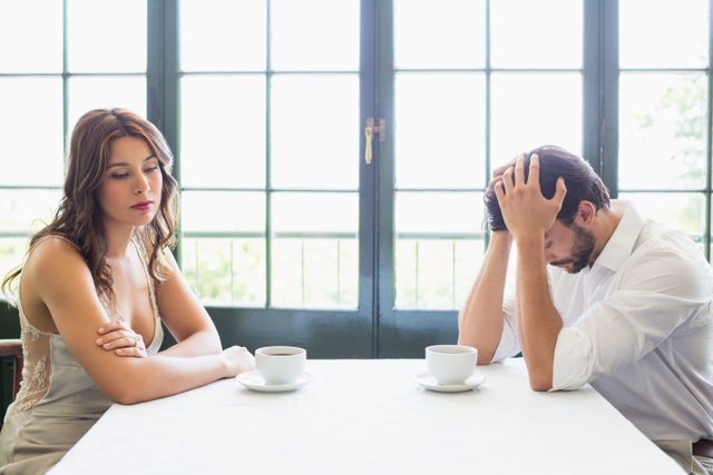 Young couple sitting at a table in a restaurant, both looking distressed and unhappy. The woman is looking down with a sad expression, while the man has his head in his hands. This image can be used to illustrate themes of relationship issues, emotional stress, and difficult conversations. Suitable for articles, blogs, or advertisements related to mental health, relationships, and counseling.