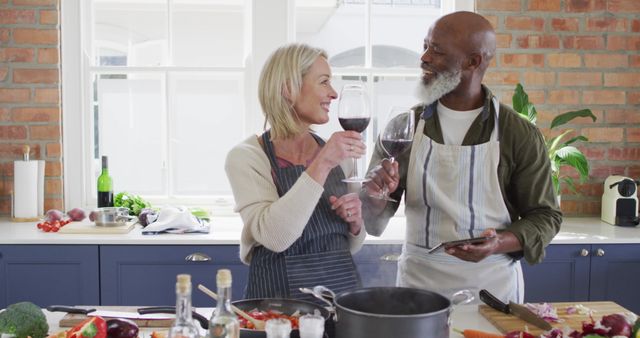 Senior couple in modern kitchen enjoying wine while cooking together. They are smiling and bonding over meal preparation. Perfect for themes about relationships, healthy lifestyles, cooking at home, and retirement happiness.