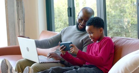 African American Man and Boy Sharing Smartphone and Laptop on Leather Sofa, Laughing Together