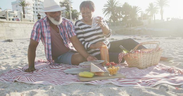 Senior couple enjoying a romantic picnic on a sunny beach. The scene exudes relaxation and happiness with a beach in the background, and the picnic setup includes a basket and various foods. Ideal for ads or articles focusing on senior living, vacation destinations, healthy living, or romantic getaways.