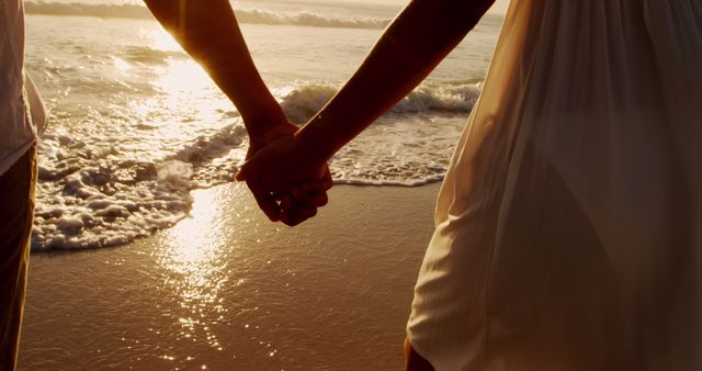 Biracial couple enjoys a romantic sunset on the beach. Their clasped hands symbolize love and companionship in a serene outdoor setting.