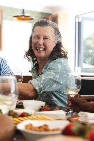 Mature female friends laughing, sharing wine at sunlit kitchen table gathering