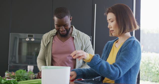 Interracial couple engaging in cooking activities in a modern kitchen environment. The man is holding a container while the woman is chopping vegetables. Suitable for use in advertisements or articles about healthy living, home cooking, multicultural relationships, or cooking tutorials. Highlights themes of teamwork, friendship, domestic activities, and healthy food preparation.