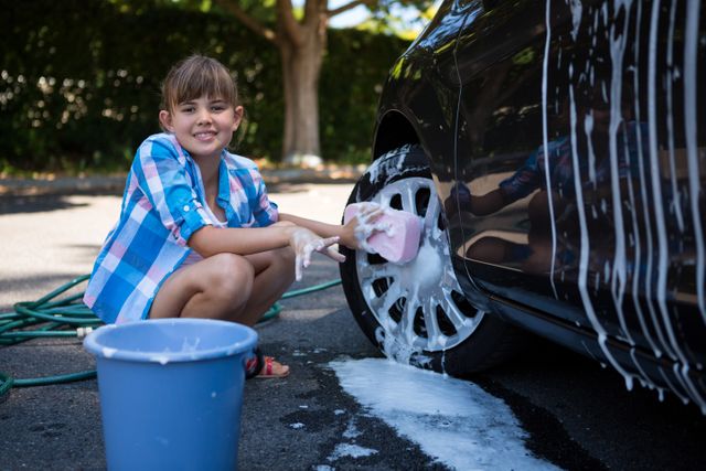Teenage girl washing a car with a sponge and soapy water on a sunny day. She is smiling and enjoying the outdoor activity. Ideal for use in advertisements for car cleaning products, family activities, summer fun, and vehicle maintenance services.