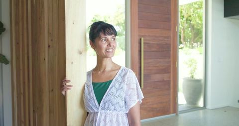 Mature woman smiling in minimalist entryway holding wooden post wearing crocheted cover-up