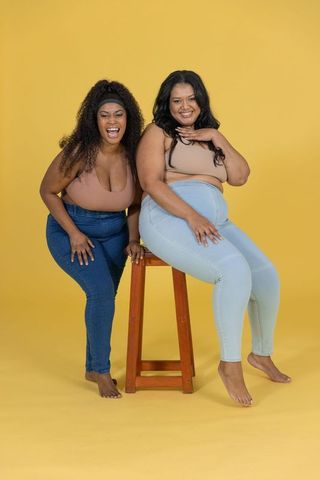 Plus-size diverse women laughing and posing on wooden stool in bright yellow studio