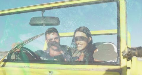 Young couple driving yellow open-top jeep through sunlit desert smiling while checking smartphone