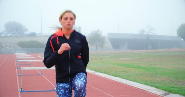 A young Caucasian woman is jogging on a track field on a foggy day, with copy space. Her focused expression and athletic attire suggest a commitment to fitness and health.