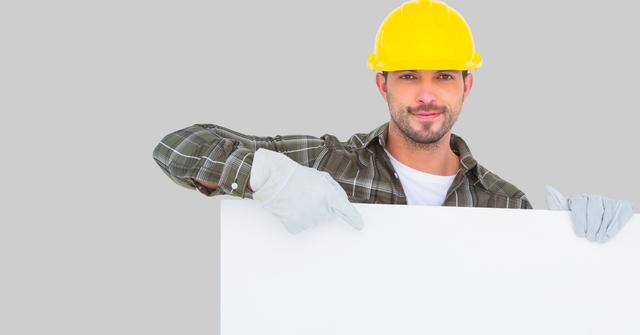 Construction worker dressed in plaid shirt, yellow hard hat, and safety gloves pointing at a blank white billboard. Ideal for advertising and marketing concepts, safety instructions, or industrial announcements. Suitable for business promotions, construction ads, or workplace safety campaigns.