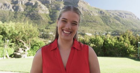 Smiling woman standing in red sleeveless blouse in sunlit garden with mountain backdrop