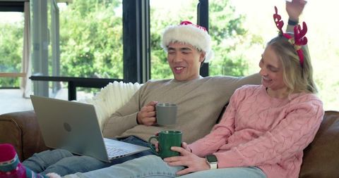 Couple relaxing on couch using laptop and drinking hot drinks in festive sweaters