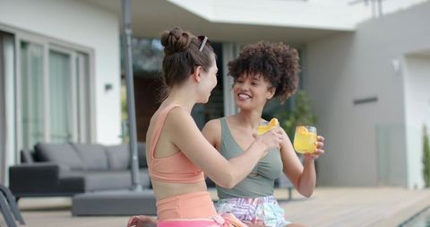 Friends toasting on terrace in activewear crop tops while sipping refreshing orange drinks