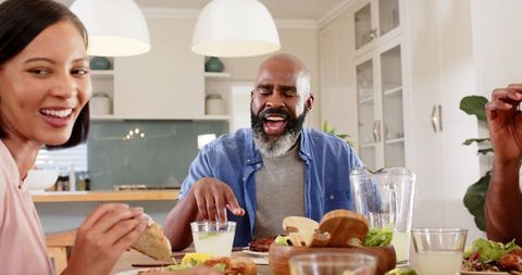 Diverse friends laughing and sharing homemade sandwiches and salad around kitchen table