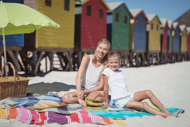 Mother and daughter enjoying a sunny day at the beach, sitting on a blanket with a picnic basket. Colorful beach huts in the background add a vibrant touch. Ideal for use in family vacation promotions, summer holiday advertisements, and lifestyle blogs.