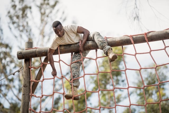 Military soldier climbing a net during an obstacle course in boot camp. The soldier is wearing a camouflage uniform and boots, demonstrating strength and determination. This image can be used for themes related to military training, physical fitness, teamwork, and overcoming challenges.