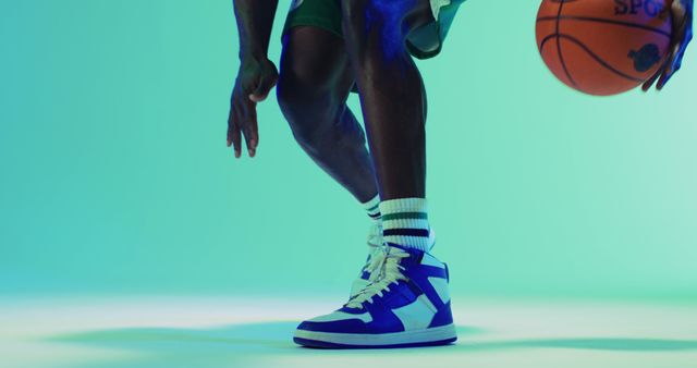This dynamic image captures the moment an athlete dribbles a basketball in a studio setting. The individual is wearing blue high-top sneakers and green shorts, emphasizing the action and intensity of the sport. Perfect for use in sports marketing materials, fitness blogs, athletic gear promotions, or basketball training programs.
