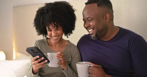 African American couple sharing coffee and laughing together in cozy bedroom