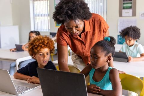 African American teacher guiding elementary students using laptops in modern classroom