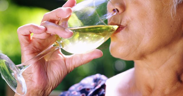 This close-up image shows an elderly woman enjoying a glass of white wine outdoors, capturing a moment of leisure and relaxation. The focus is on her face and hand, highlighting her savoring the drink. This image can be used in contexts promoting senior lifestyle content, wine and beverage advertisements, health and wellness articles, and outdoor leisure activities marketing.