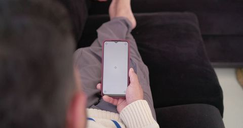 Indian man sitting on sofa holding smartphone with blank screen for app mockup