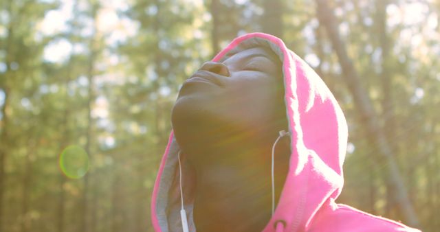 Person wearing a pink hoodie standing in a forest with sunlight filtering through the trees, eyes closed, and face upturned, exuding relaxation and serenity. Headphones hint musical enjoyment or mindfulness practice. Useful for lifestyle blogs, meditation apps, wellness articles, and nature experiences promotions.