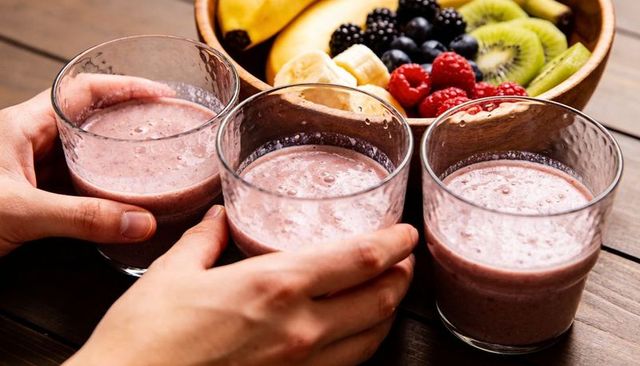 Hands holding three pink berry smoothies with fresh fruit bowl on rustic wooden table