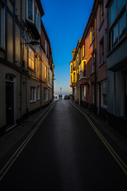 Narrow urban street bathed in evening light with vivid building facades leading to sea in distance. Ideal for travel blogs, architectural studies, urban photography collections, and coastal nostalgia themes.