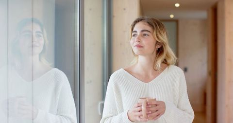 Serene woman holding mug gazing out glass door in minimalist home