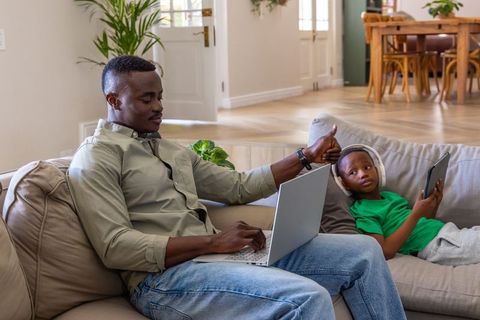 African american father giving thumbs up while working on laptop with son holding tablet