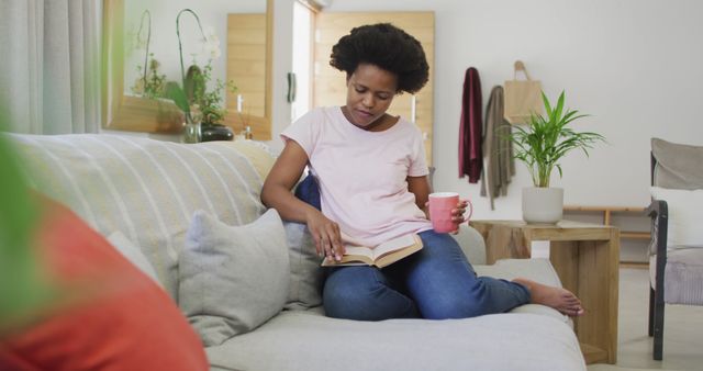 Woman is sitting on comfortable couch in cozy living room, casually dressed in jeans and a t-shirt, holding coffee mug while reading a book. Houseplants and soft furnishings create a warm and inviting atmosphere, making it perfect for themes of relaxation, leisure, self-care, and home life. Ideal for promoting home decor, lifestyle blogs, and articles on creating a relaxing home environment.