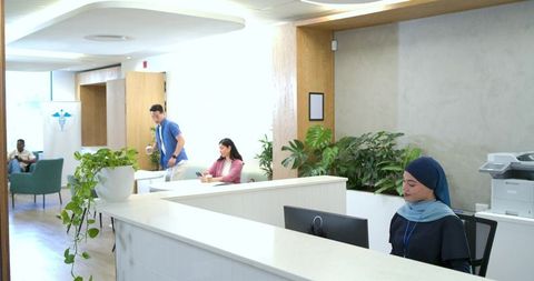 Receptionist wearing blue hijab at modern inclusive medical front desk with greenery