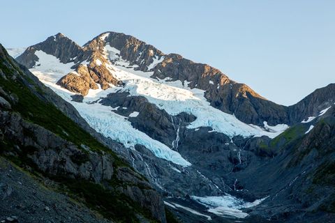 Sunlit Alpine Glacier Flowing Through Rugged Rocky Peaks Under Clear Blue Sky at Dawn