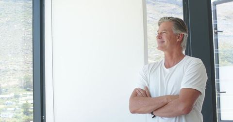 Senior man standing with arms crossed looking out window in minimalist modern living space