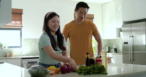 Smiling asian couple preparing healthy meal at kitchen island with fresh produce and herbs