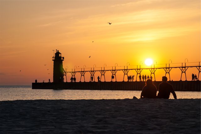 Couple sitting on beach watching sunset, with lighthouse and pier in background. People strolling along pier, birds flying across sky. Ideal for themes like romance, travel brochures, relaxation, scenic beauty, or beach vacations.