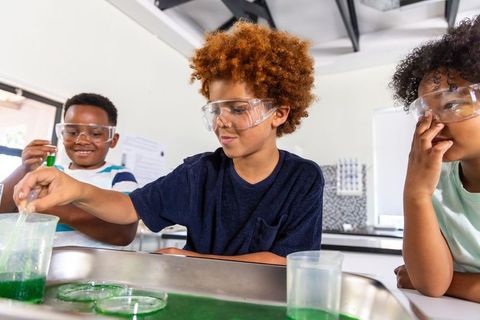 African american kids wearing safety goggles mixing green solution in classroom lab stem