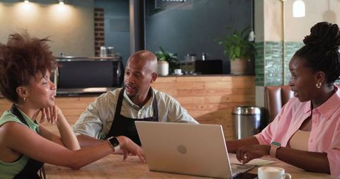 Three cafe staff planning shift around laptop in cozy coffeehouse with warm wooden interior
