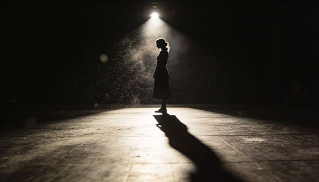 Standing silhouette under spotlight on empty stage casting long dramatic shadow and dust