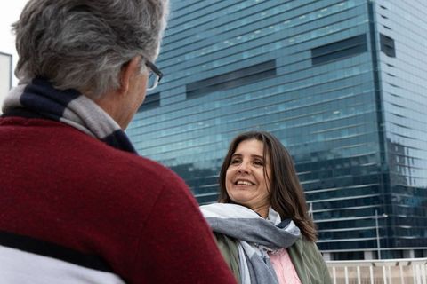 Mature couple enjoying rooftop conversation near glass tower, smiling woman wearing scarf