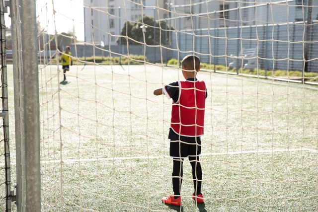 Two boys soccer players practicing on a green football pitch on a sunny day, goalkeeper standing in goal, seen through net. Childhood healthy lifestyle competition.