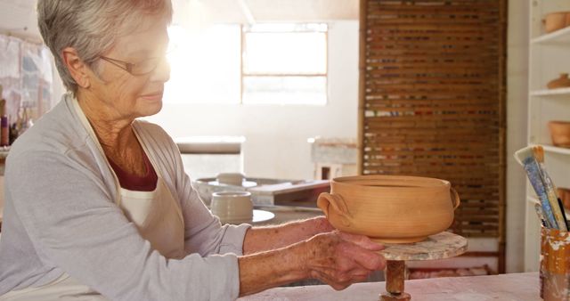 Elderly woman shaping clay pottery in brightly lit art studio. Ideal for content on hobbies, retirement activities, creative arts, senior lifestyle, or artistic workshops.