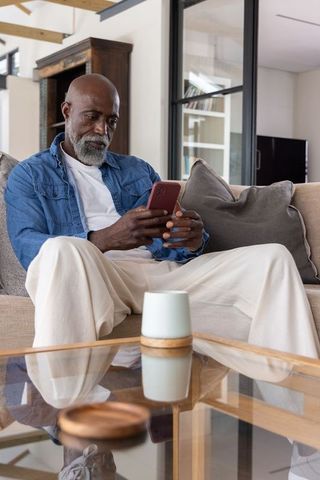 Mature african man relaxing on sofa reading smartphone with red phone wearing denim shirt in modern