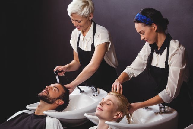 Two professional hairdressers are washing the hair of a male and a female customer at a salon. The customers are reclining comfortably at the wash stations, enjoying a relaxing hair care treatment. This image is ideal for use in advertisements for beauty salons, hair care products, or articles about professional hair care services.