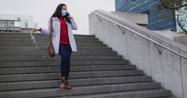 Woman wearing protective face mask, talking on smartphone while walking downstairs in urban setting. Ideal for use in articles or content related to pandemic safety measures, outdoor activities, urban lifestyle, and public health.
