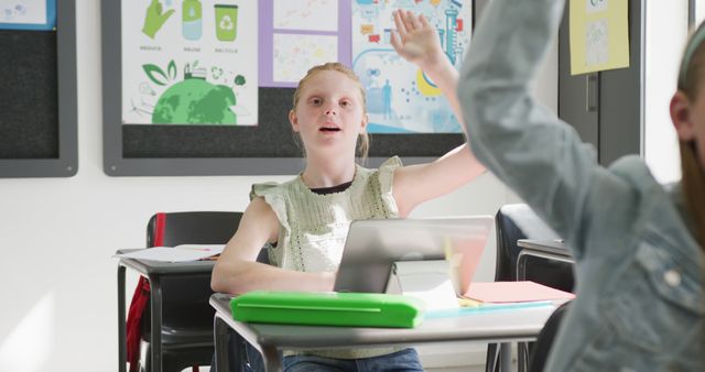 Girl in a classroom setting actively participating by raising her hand. She is seated at her desk with a tablet in front of her, suggesting a modern, technology-enabled learning environment. Suitable for use in educational content, articles on classroom engagement, and promotions for educational technology and school programs.