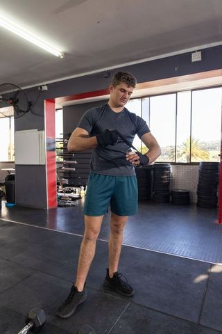 Young man fastening wrist wraps before dumbbell workout in sunlit commercial gym