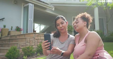 Two diverse women sharing smartphone on sunny suburban lawn, relaxing on porch steps, smiling