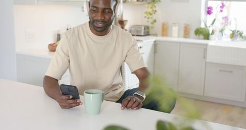 Man listening with headphones holding smartphone drinking from green mug in bright kitchen
