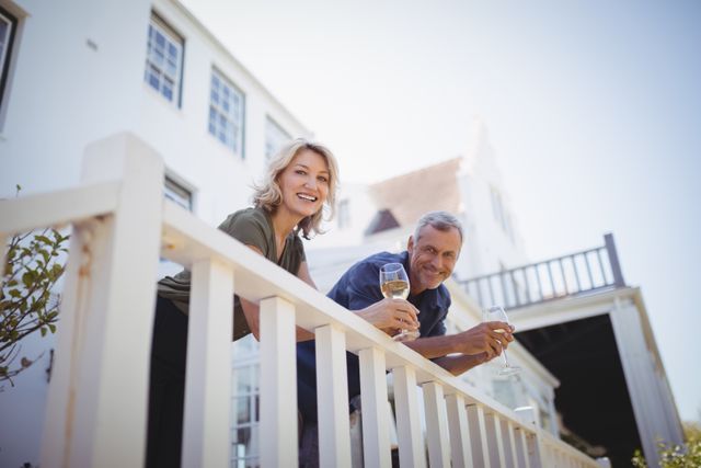 Mature couple smiling and enjoying glasses of wine on a balcony at home. Ideal for use in advertisements or articles about lifestyle, relaxation, home living, and senior relationships. Perfect for promoting wine brands, retirement communities, or home improvement services.